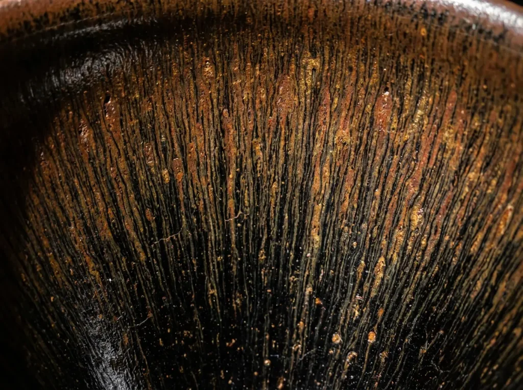 Close-up macro photography of Tenmoku tea cup showing hare's fur glaze pattern with golden brown vertical streaks on black ceramic surface
