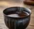 Authentic Tenmoku tea cup featuring iron-rich black glaze with silver hare's fur patterns, filled with amber pu-erh tea on a wooden table
