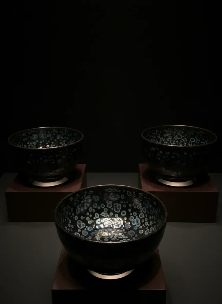 How Tenmoku Became Popular in Japan: The Zen Tea Connection 4 Three National Treasure yohen tenmoku tea bowls displaying shimmering iridescent oil-spot glaze with blue and purple light reflections on dark ceramic surface, museum exhibition