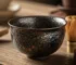 Close-up of a Tenmoku tea cup with dark oil spot glaze pattern showing metallic iron crystals on a wooden tea table